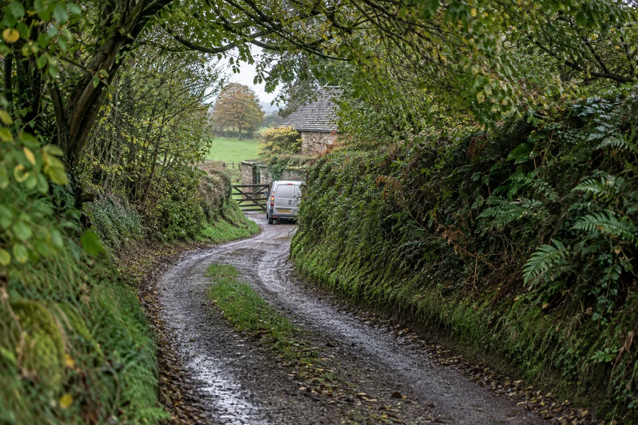 Chemin rural étroit et humide entre des haies, avec une camionnette près du portail.